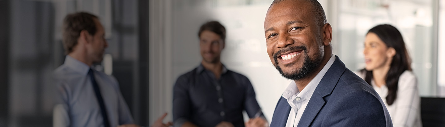 Imagem de um executivo negro olhando para a câmera e sorrindo. Ao fundo, três pessoas em desfoque conversando